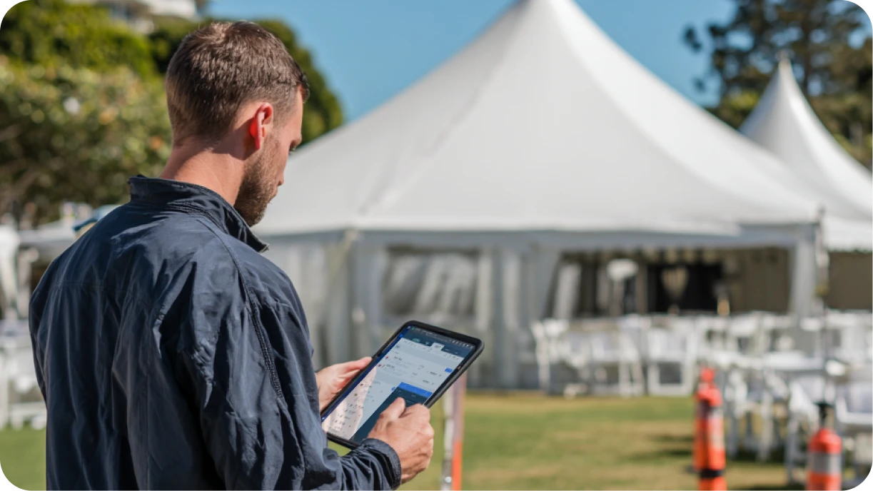 Event rental coordinator using Goodshuffle Pro software on tablet while overseeing tent and lighting setup at venue