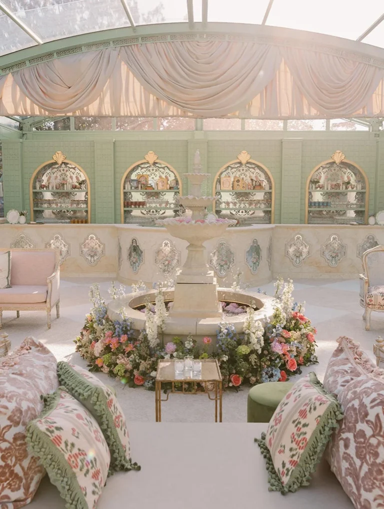 A European garden-inspired tent reception with a tiered stone fountain ringed by colorful florals, draped ceiling, and vintage lounge seating