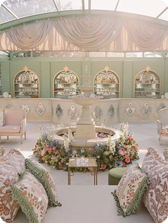 A European garden-inspired tent reception with a tiered stone fountain ringed by colorful florals, draped ceiling, and vintage lounge seating