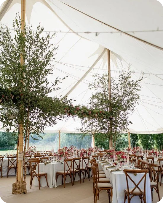 A sailcloth marquee wedding tent with living birch trees as center poles joined by a suspended garland of greenery and pink florals, above long banquet tables set with taper candles and wooden crossback chairs.