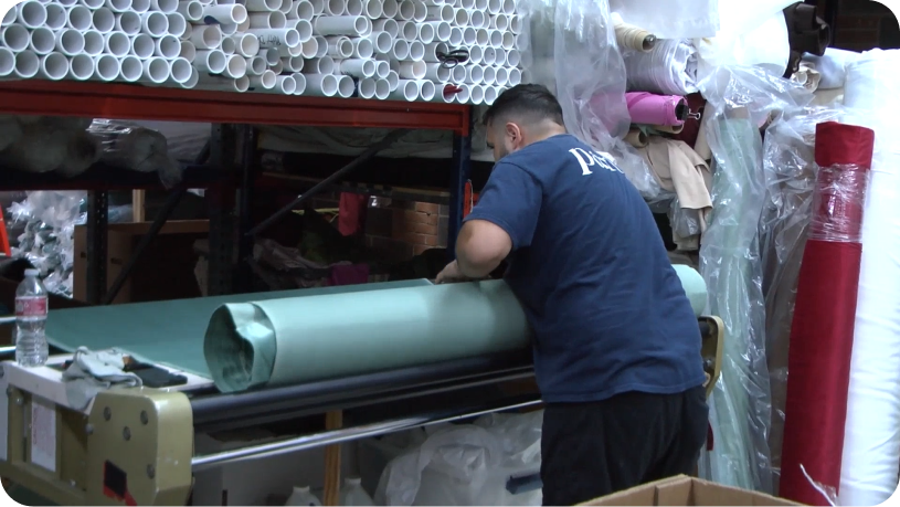 Crew member rolling a drape panel on a fabric roller machine, with PVC roll cores racked above