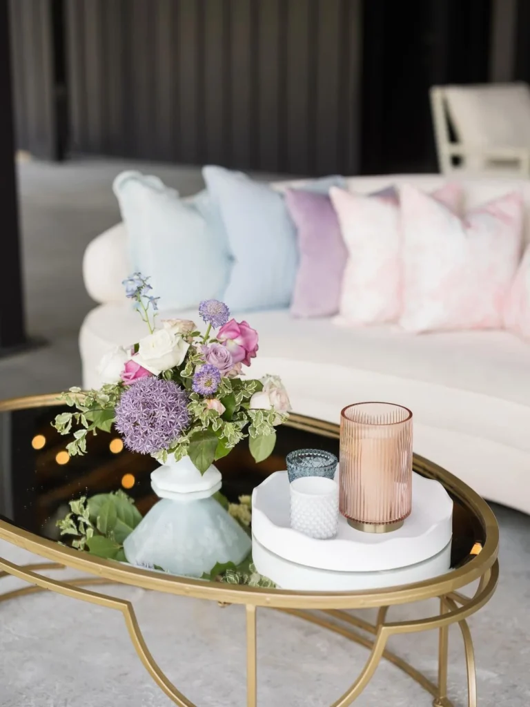 A pastel floral centerpiece of roses, allium, and delphinium on a gold-framed glass coffee table, beside a cream curved sofa layered with blue, lavender, and blush pillows.