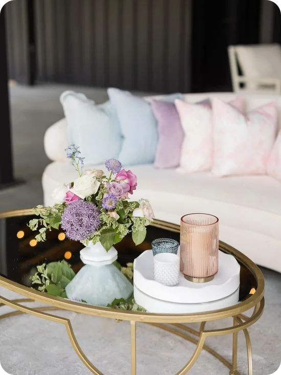 Un centro de mesa floral en tonos pastel compuesto por rosas, allium y delfinios, sobre una mesa de centro de vidrio con marco dorado, junto a un sofá curvo color crema adornado con cojines azules, lavanda y rosa pálido.