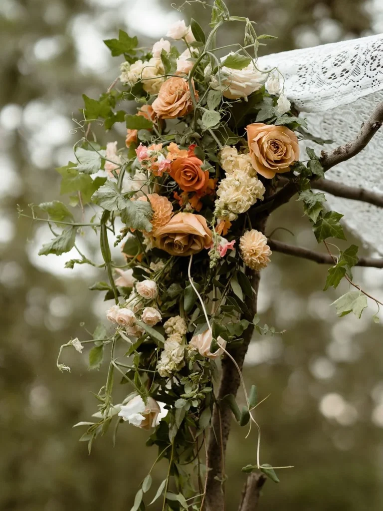 A boho wedding chuppah adorned with peach and terracotta roses winding up birchwood posts, trailing greenery, and draped lace, set among pine trees.
