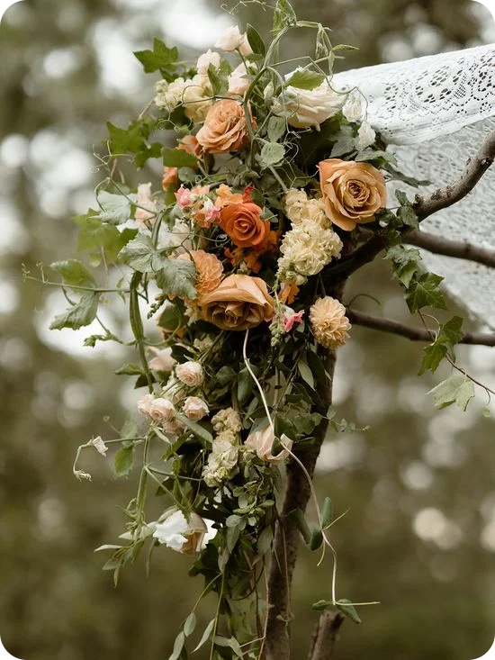 A boho wedding chuppah adorned with peach and terracotta roses winding up birchwood posts, trailing greenery, and draped lace, set among pine trees.