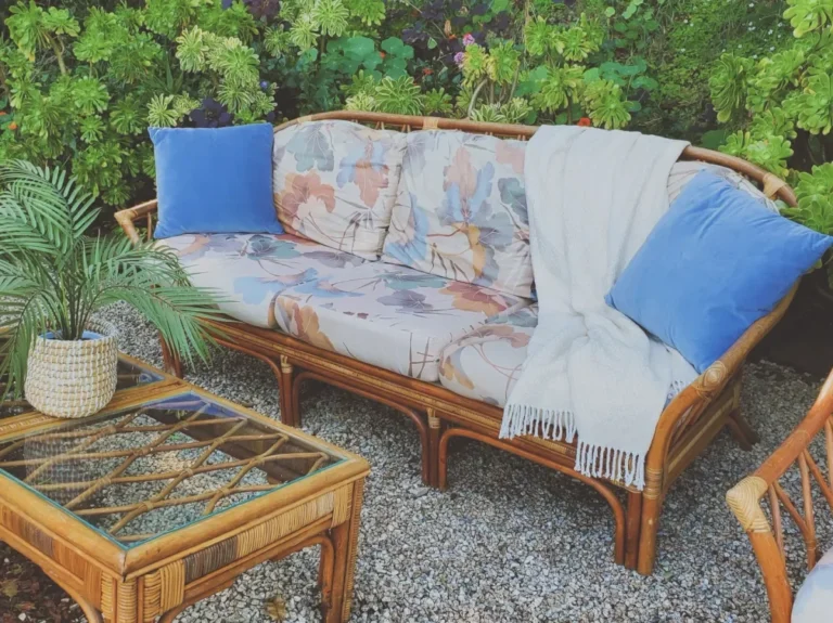 A rattan sofa with floral-print cushions and blue velvet pillows beside a glass-top rattan coffee table in a lush garden lounge setup