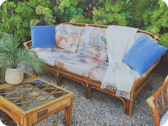 Un sofá de ratán con cojines de estampado floral y almohadas de terciopelo azul junto a una mesa de centro de ratán con tapa de vidrio, en un exuberante rincón de descanso al aire libre
