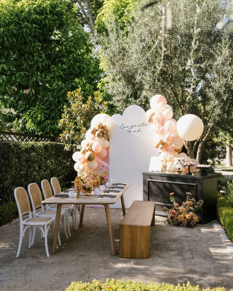 Decoración de mesa para una fiesta de graduación al aire libre con una guirnalda de globos color rosa pálido, un fondo con el mensaje «Felicidades, graduado», una mesa de madera y una barra de postres
