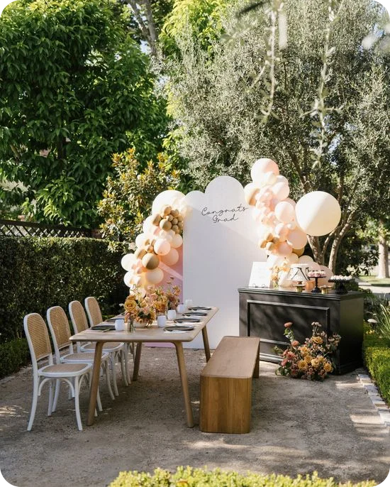 Decoración de mesa para una fiesta de graduación al aire libre con una guirnalda de globos color rosa pálido, un fondo con el mensaje «Felicidades, graduado», una mesa de madera y una barra de postres