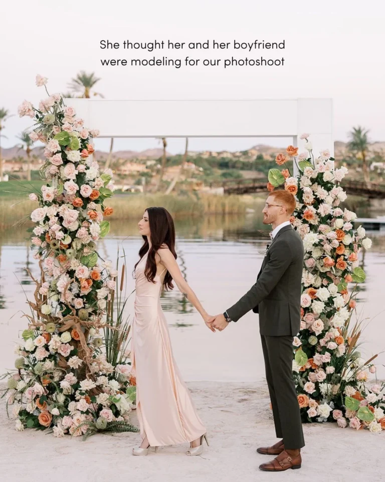 Couple holds hands between two lush floral arch pillars at a lakeside surprise proposal