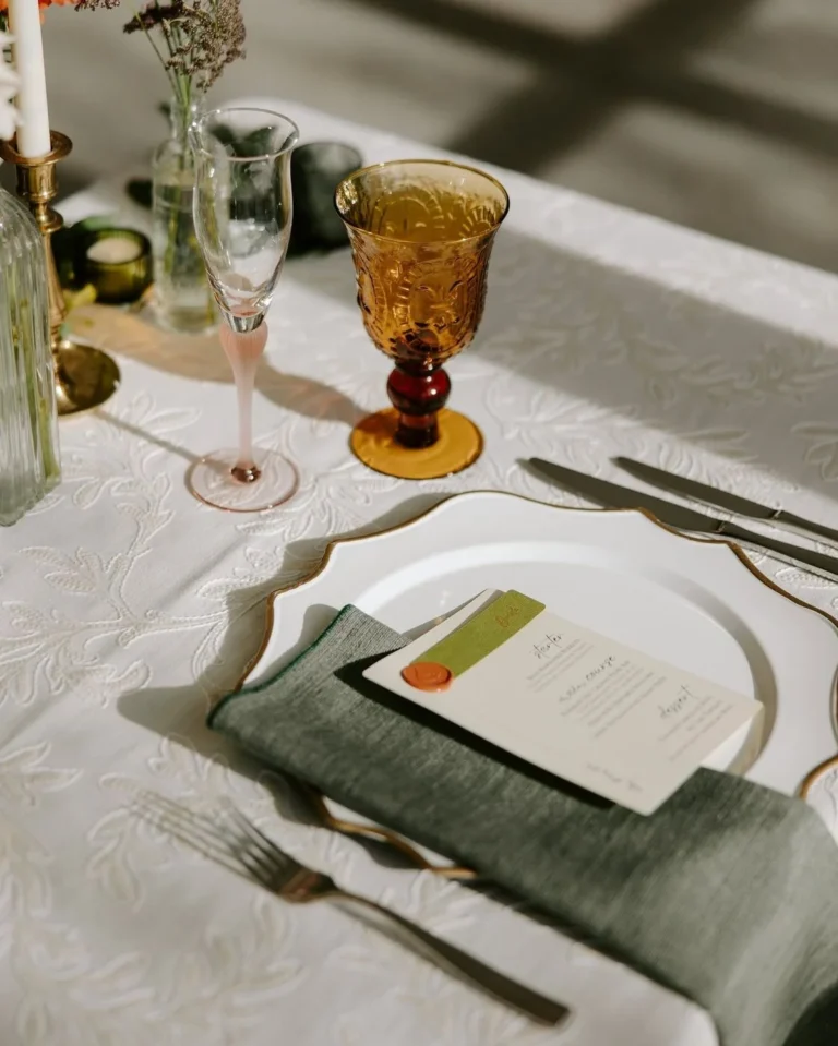 Une décoration de table composée d'une assiette de présentation festonnée à bord doré, d'une serviette vert sauge, d'une carte de menu scellée à la cire et d'un gobelet ambré.