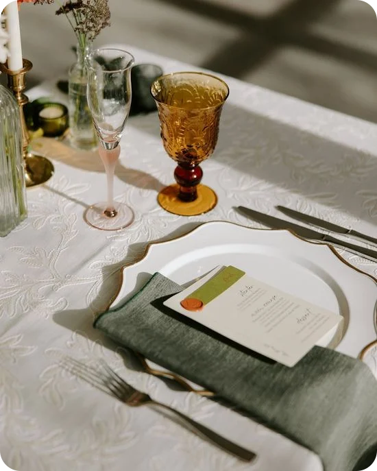 Une décoration de table composée d'une assiette de présentation festonnée à bord doré, d'une serviette vert sauge, d'une carte de menu scellée à la cire et d'un gobelet ambré.