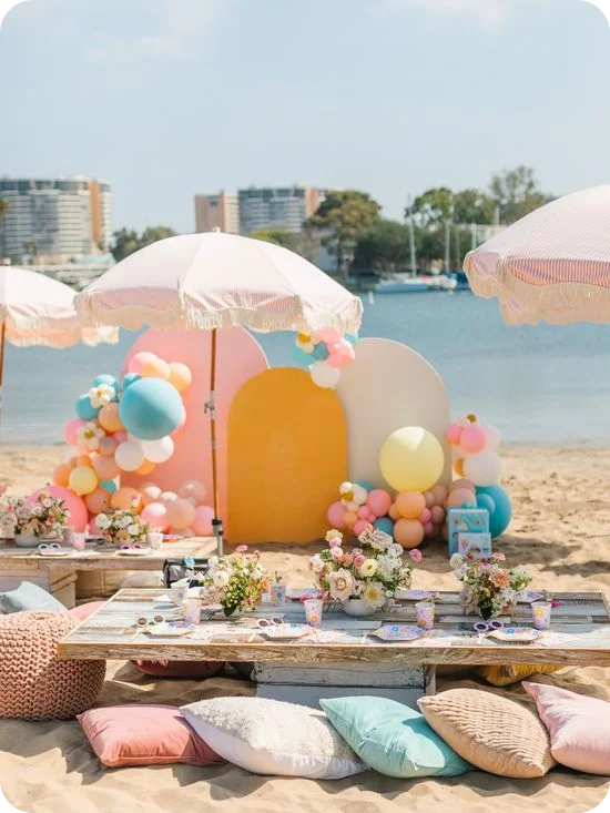 Fiesta de picnic en la playa en tonos pastel, con mesitas bajas de madera, sombrillas con flecos, guirnaldas de globos y cojines en la arena