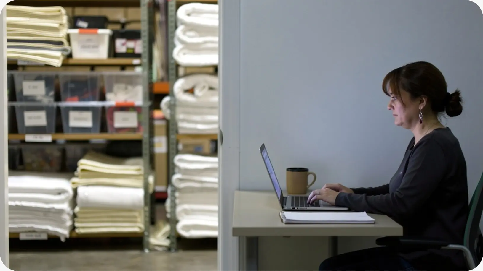 La dueña de una empresa de alquiler de artículos para eventos está sentada en un escritorio en la oficina de su almacén, revisando su catálogo de alquiler en línea en una computadora portátil, con estantes llenos de ropa de cama doblada y cajas de almacenamiento detrás de ella.