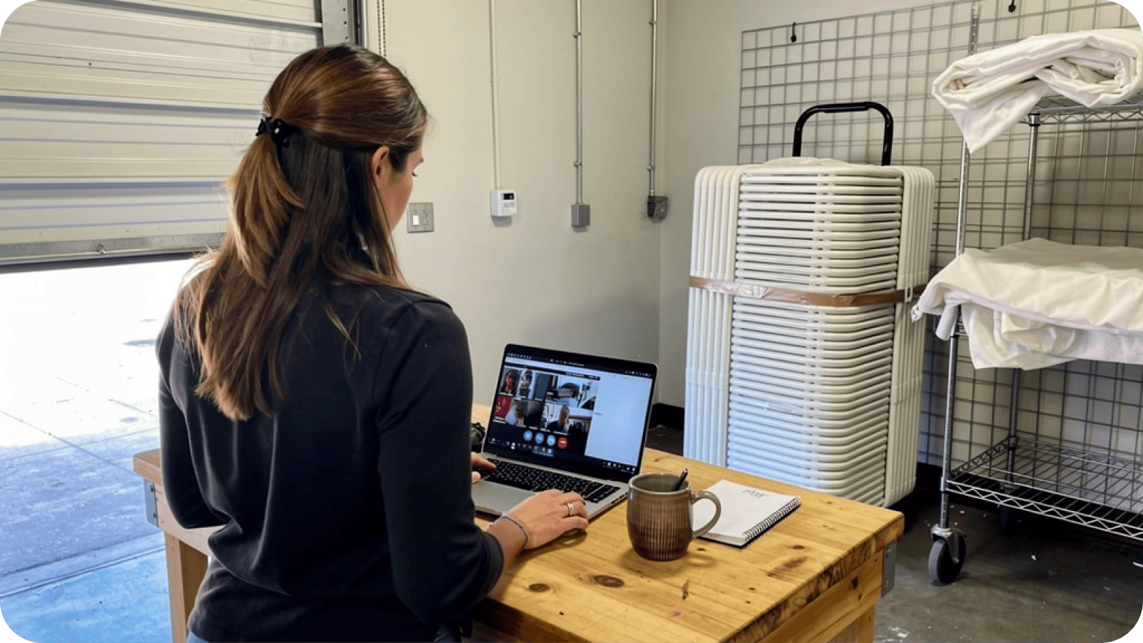 Event rental owner at a warehouse workbench watching a recorded Summit session on her laptop, with a strapped stack of folding chairs on a hand truck and linens racked behind her.
