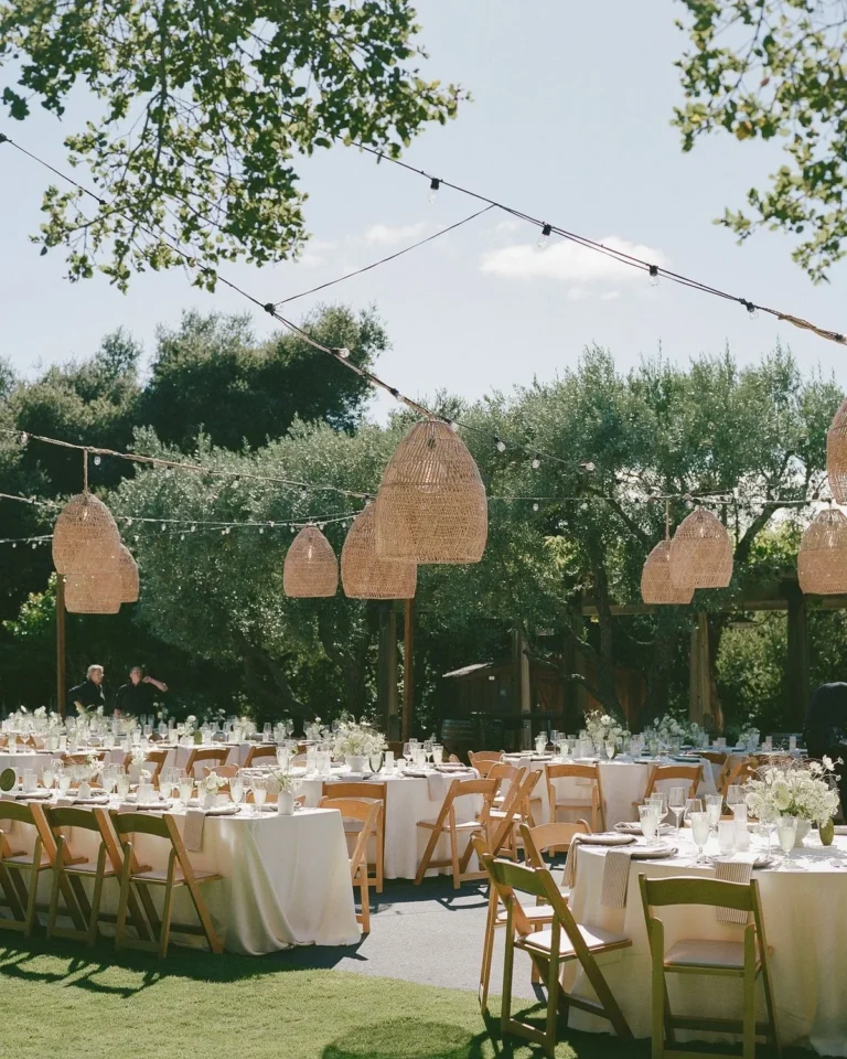 Rattan pendant lanterns and string lights over an outdoor wedding reception with white linens and wooden folding chairs