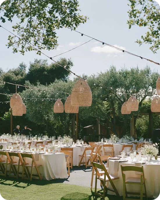 Rattan pendant lanterns and string lights over an outdoor wedding reception with white linens and wooden folding chairs