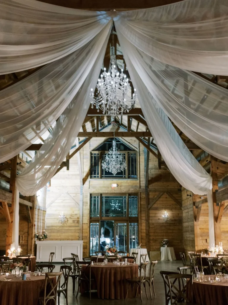 Telas blancas drapeadas y candelabros de cristal sobre mesas redondas de cena en una recepción de boda celebrada en un granero.
