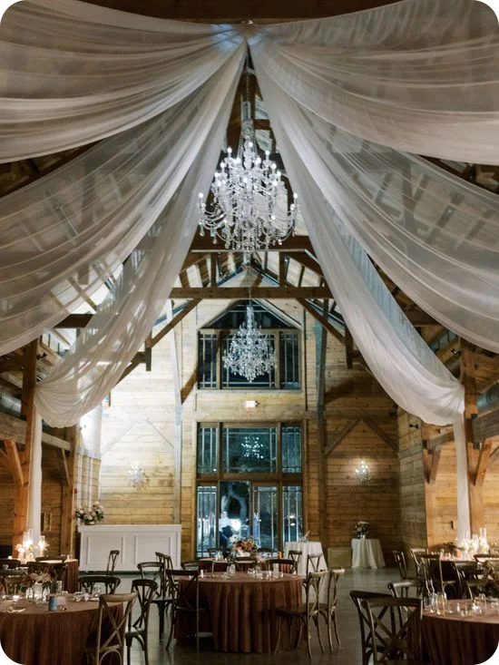 Telas blancas drapeadas y candelabros de cristal sobre mesas redondas de cena en una recepción de boda celebrada en un granero.