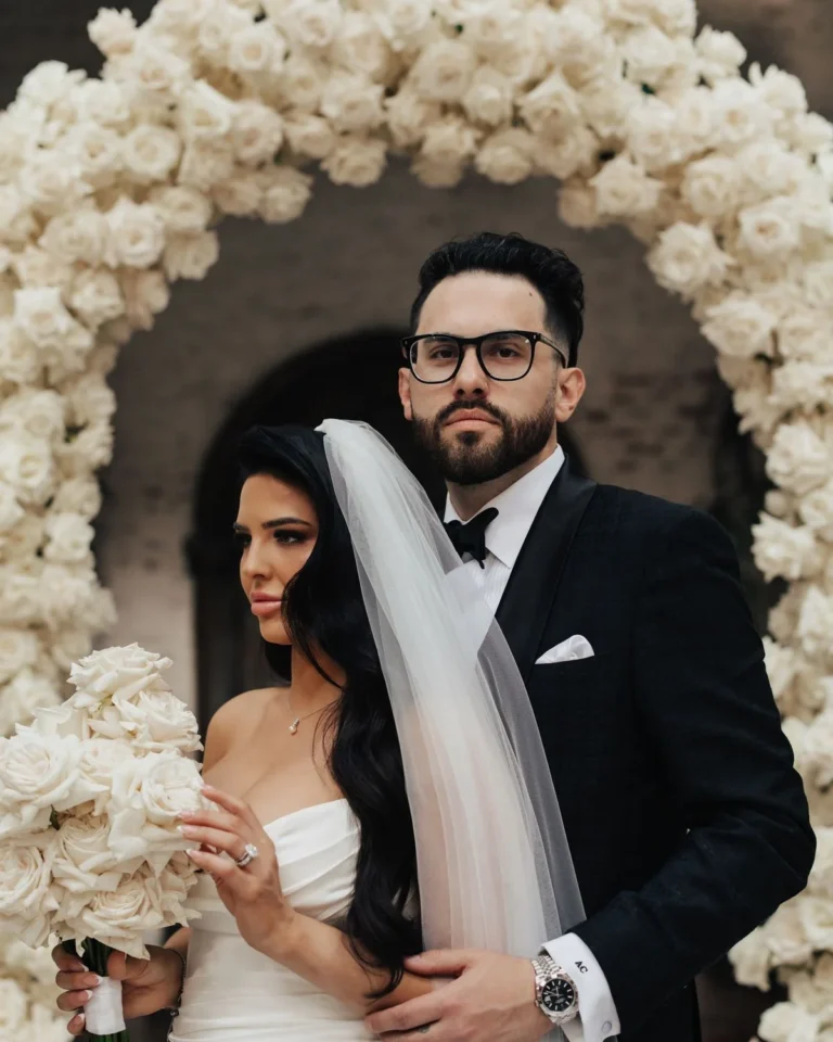 Les mariés se tiennent devant une arche de cérémonie ornée de roses blanches ; la mariée tient un bouquet de roses ivoire assorti.