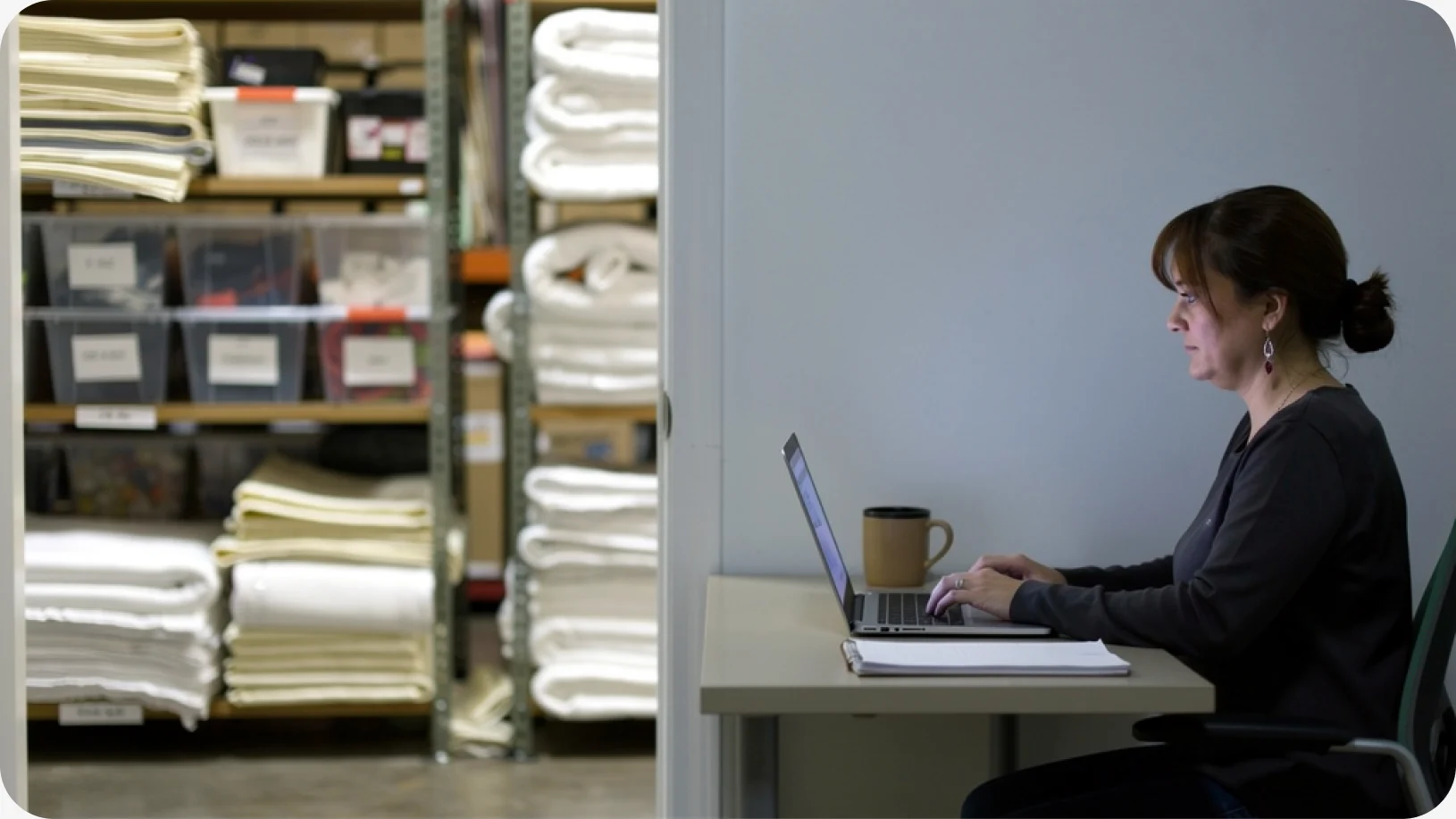 La dueña de una empresa de alquiler de artículos para eventos está sentada en un escritorio en la oficina de su almacén, revisando su catálogo de alquiler en línea en una computadora portátil, con estantes llenos de ropa de cama doblada y cajas de almacenamiento detrás de ella.