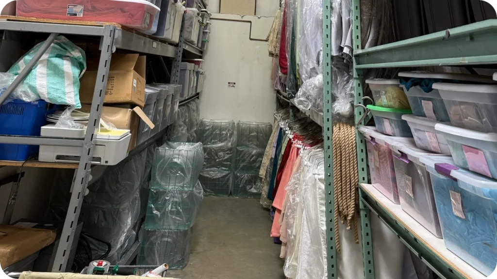 A narrow storage aisle in Waterford Event Rentals' warehouse, with boxes and lidded bins on the shelves and linens hanging in plastic along one side.