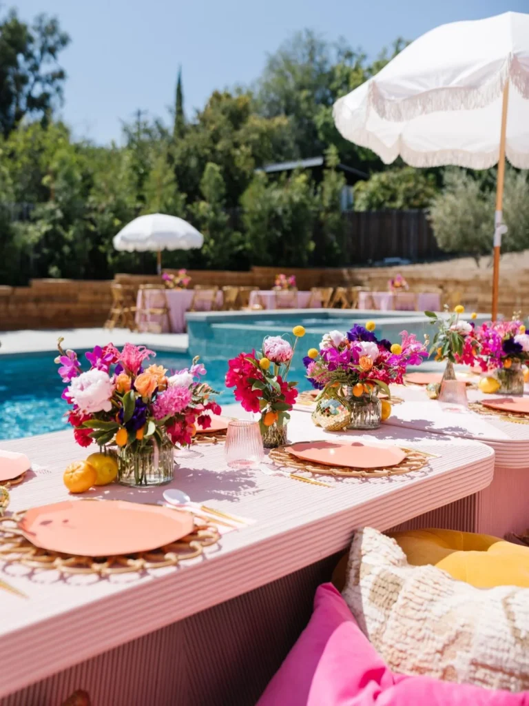 Table de réception en plein air au bord de la piscine, de couleur rose, avec des assiettes corail, des sous-assiettes en rotin, des centres de table floraux aux couleurs vives et des parasols à franges