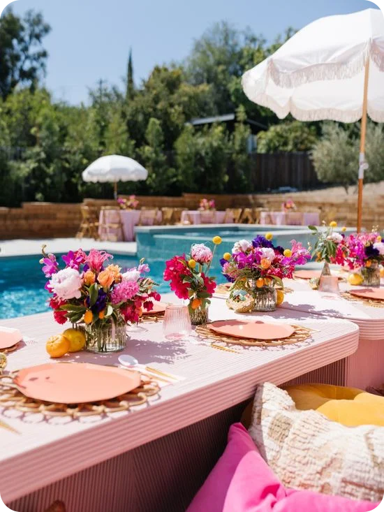Table de réception en plein air au bord de la piscine, de couleur rose, avec des assiettes corail, des sous-assiettes en rotin, des centres de table floraux aux couleurs vives et des parasols à franges